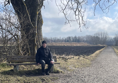 Mann sitzt auf einer Bank an einem Feld und schaut Richtung Kamera. Als Vorlagenbild ist das Motiv zu weit entfernt